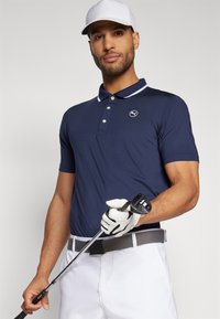 Polo shirt bleu marine avec col blanc, logo sur la poitrine gauche, associé à un pantalon blanc et une ceinture grise. Le mannequin tient un club de golf avec un gant à une main.