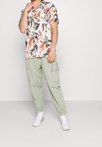 Chemise à manches courtes à motif floral en blanc, avec des touches vives de vert, rose et orange. Associée à un pantalon cargo vert clair et des baskets blanches.