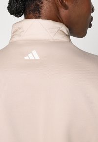 Béžová bunda s prošívaným límcem, na zadní straně je bílé logo Adidas. Tkanina vypadá hladce s jemnou strukturou.