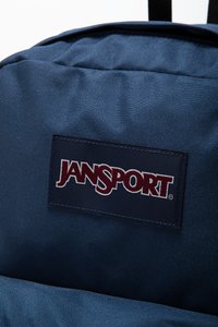 Marineblå stoffryggsekk med en teksturert overflate og et sydd Jansport-logo-patch i burgunder og hvitt. Justerbare svarte stropper.