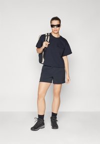 Marinblå kortärmad t-shirt, matchande shorts, robusta svarta vandringskängor och solglasögon; bär en svart ryggsäck med beige axelremmar.