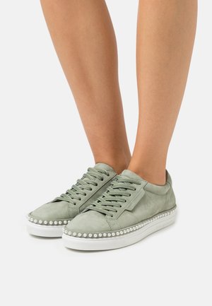 Osoba w jasnozielonych sneakersach z białymi podeszwami i perłowymi ćwiekami wzdłuż krawędzi, stojąca na jednolitym białym tle.