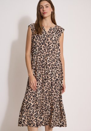 Kvinde iført ærmeløs beige midikjole med sort og brun leopardprint, stående mod en ensfarvet lys baggrund.