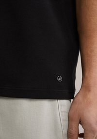 Černé bavlněné tričko s hladkým povrchem, na spodním lemu se malým stříbrným logem. V kombinaci se světle zbarvenými kalhotami.