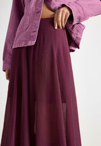 Person iført et gennemsigtigt blommefarvet skørt og en lyserød denimjakke, med den ene hånd hvilende på taljen over skørtet.