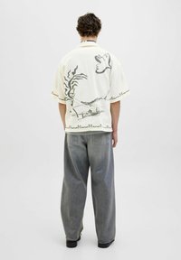 Chemise à manches courtes de couleur crème avec un design graphique noir, associée à un pantalon en denim gris à coupe décontractée. Dispose d'un col relaxé et d'un détail au dos.