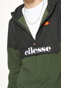 Zielono-czarna bluza polarowa z półzamkiem, z białym logo "ellesse" oraz wyszywanym logo w górnym lewym rogu.