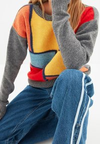 Bunter Strickpullover mit einem geometrischen Muster in orange, gelb, blau und roten Abschnitten. Graue Ärmel und gerippter Ausschnitt sind ebenfalls erwähnt.