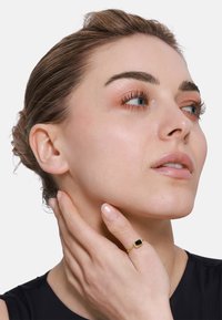 Anello d'oro con una pietra nera rettangolare, indossato su una mano che tocca la mascella. Il modello ha un trucco neutro e i capelli raccolti in uno chignon.