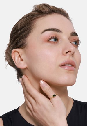 Goldring mit einem rechteckigen schwarzen Stein, der an einer Hand getragen wird, die den Kiefer berührt. Das Model hat ein neutrales Make-up und die Haare zu einem Dutt frisiert.