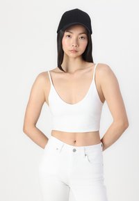 Bijeli crop top s dubokim V-izrezom i tankim naramenicama, uparen s visokim bijelim trapericama i crnom bejzbol kapom. Glatka tekstura tkanine.