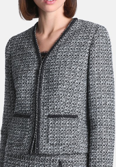 Femme portant une veste en tweed noir et blanc avec de petites poches avant et un col en V laissant apparaître un vêtement en dentelle en dessous.
