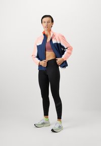 Chaqueta deportiva en azul marino con mangas de color rosa claro, leggins negros de cintura alta y zapatillas de correr gris verdoso con acentos en rosa.