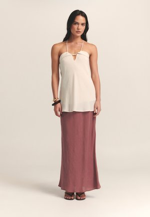 Bezrukava kremasto bijela bluzica s detaljem u obliku ključa, u kombinaciji s dugom, lepršavom ljubičastom suknjom. Glatka tekstura, minimalistički dizajn i crne sandale.