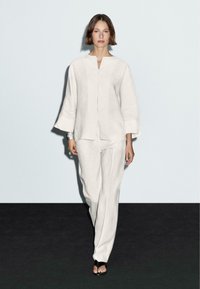 Tenue en lin légère de couleur crème, composée d'une chemise à boutons, à manches longues, et de pantalons larges, sur un fond neutre.