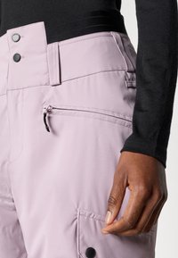 Lyserøde skibukser med en tekstureret overflade, der har en lynlåslomme, knaplukninger og en sort talje.