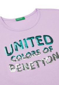 Camicia lilla con etichetta verde e testo "UNITED COLORS OF BENETTON" in paillettes turchesi e argento sul davanti.