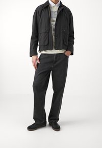 Homme portant une veste noire sur un polo rayé, un jean noir ample et des baskets noires, se tenant avec une main dans sa poche.