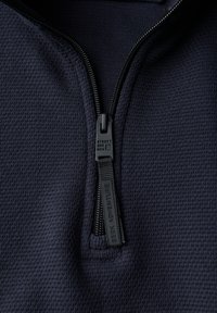 Detailní záběr na tmavě modrou texturovanou látkovou bundu s černým táhlem zipu označeným "HLEDEJ DOBRODRUŽSTVÍ" a "STREET ONE MEN".