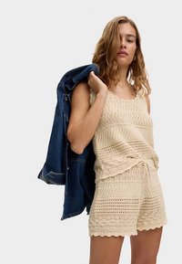 Stickad linne i beige med ett texturerat mönster, kombinerad med matchande shorts. En jeansjacka hänger över ena axeln.