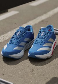 Ζευγάρι μπλε παπούτσια για τρέξιμο Adidas Adizero με λευκές και κόκκινες λεπτομέρειες τοποθετημένα σε υφή εξωτερικής επιφάνειας.