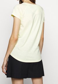 Gul bomulds T-shirt med korte ærmer, buet kant og afslappet pasform. Båret sammen med sorte shorts; minimal syning synlig.