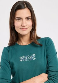 Teal Langarmshirt mit weißer Blumenstickerei auf der Brust, aus weichem Material mit rundem Halsausschnitt und taillierter Passform.