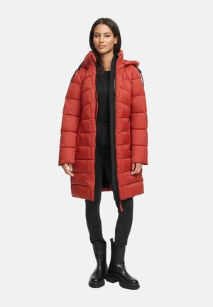 Woman wearing a long red quilted puffer coat over black top, black jeans, and black ankle boots, standing against a plain background.