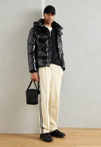 Giacca puffer nera lucida con cappuccio, abbinata a una maglietta nera a maniche lunghe e pantaloni della tuta beige con strisce laterali; tiene in mano una piccola borsa nera.