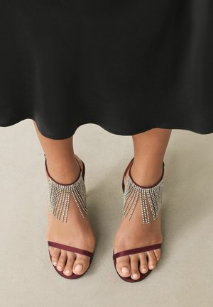 Burgunderfarbene Sandalen mit einem Riemen-Design, mit schimmernden silbernen Kettenakzenten, die vom Knöchel herabfallen. Glatte matte Textur.