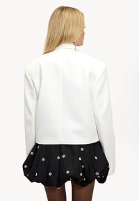 Witte blazer met een gestructureerd silhouet, met scherpe schouders en een gladde textuur. Gecombineerd met een zwarte rok versierd met bloemmotieven.
