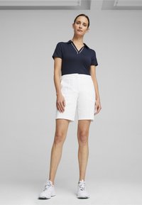 Camisa polo navy con escote en V y ribetes blancos, combinada con pantalones cortos blancos hasta la rodilla. El modelo lleva zapatos deportivos blancos.