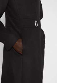 Cappotto di lana nera con una texture liscia, maniche lunghe e una fibbia rettangolare argento in vita. Tasche laterali visibili.