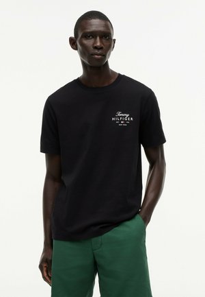 Junger Mann trägt ein schwarzes Tommy Hilfiger T-Shirt und grüne Shorts, steht mit einer Hand in der Tasche vor einem schlichten hellen Hintergrund.