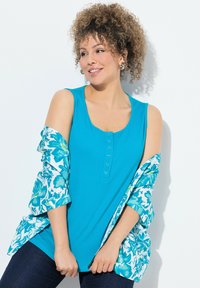 Canotta turchese con scollo rotondo e dettagli con bottoni, sovrapposta a una camicia aperta con stampa floreale, caratterizzata da tonalità di verde e blu.