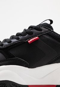 Černá atletická obuv s texturovaným černým svrškem, bílou podrážkou, červenou označovací etiketou a černými tkaničkami. Vyznačuje se gumovou podrážkou a odolným materiálem.