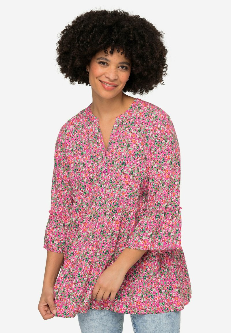 Blouse florale rose avec un décolleté en V, des manches volantes et une coupe ample. Présente de petits motifs de fleurs multicolores sur un fond clair.