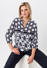 Chemisier à motif géométrique noir et blanc avec une patte de boutonnage demi-longue, des manches trois-quarts et des poignets légèrement froncés, associé à un pantalon sombre.