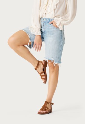 Kobieta ubrana w jasnoniebieskie, podarte szorty z denimu, białą bluzkę z falbanami oraz brązowe, skórzane sandały na płaskiej podeszwie, stojąca na jednej nodze.