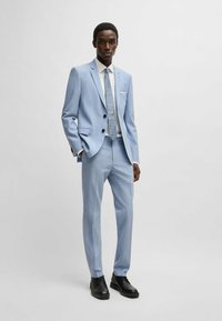 Completo blu chiaro con righe sottili, design a un petto, bottoni neri, camicia bianca e cravatta a fantasia. Scarpe nere completano il look.
