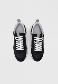 Čierne tenisky so zmesou hladkých a textúrovaných materiálov, bielymi šnúrkami a hrubou bielou podrážkou s malými vzormi na bežeckej ploche.