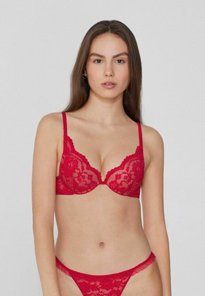 Soutien-gorge en dentelle rouge avec rembourrage, présentant des bords festonnés et des bretelles ajustables. Associé à un string en dentelle rouge, mettant en valeur des motifs floraux complexes.