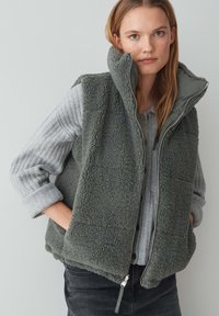 Fleece-Weste in dunkelgrün mit hohem Kragen, Reißverschluss und strukturiertem Stoff. Kombiniert mit einem hellgrauen, gerippten Langarmoberteil.
