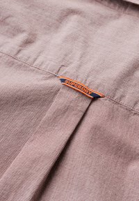 Brązowy materiał w wzór z teksturowanym splotem, z niebieską i pomarańczową etykietą "SUPERDRY" blisko szwu.