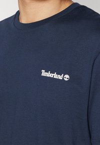 Námořnická modrá tričko s krátkým rukávem vyrobené z bavlny. Na levé části hrudi se nachází bílé logo "Timberland". Kulatý výstřih.