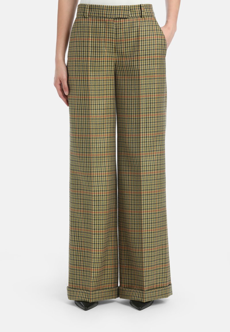 Brede broek in een groen, geel en oranje ruitpatroon. Beschikt over zijzakken en een opgerolde zoom met zwarte, puntige schoenen.
