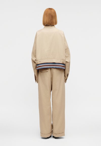 Μπεζ κοντό σακάκι με πολύχρωμη ριγέ τελική επένδυση, συνδυασμένο με μπεζ παντελόνια φαρδιάς γραμμής. Το υλικό φαίνεται λείο με ελαφριά γυαλάδα.