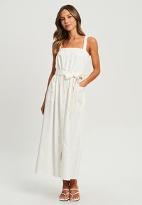 White maxi dress with adjustable straps, gathered neckline, front buttons, waist tie, and side pockets. Smooth fabric with a light texture.