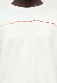 Κοντινό πλάνο ατόμου που φορά λευκό πουκάμισο με πορτοκαλί κυματιστή γραμμή στο στήθος και μικρό ανάγλυφο λογότυπο στη δεξιά πλευρά.