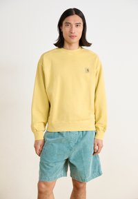 Sweatshirt jaune clair avec des poignets et un ourlet côtelés, arborant un petit logo sur la poitrine, associé à un short teal en tissu doux.